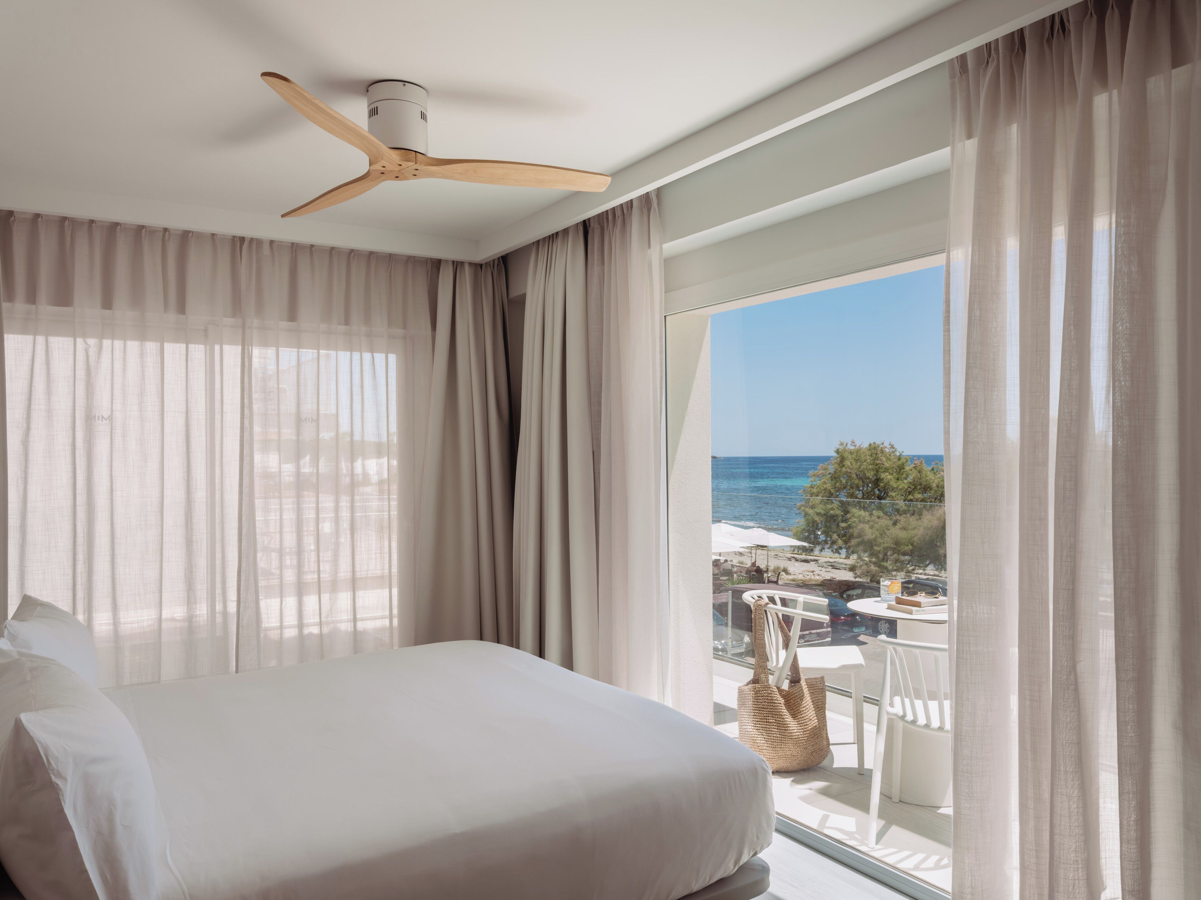 A hotel room with a bed, a ceiling fan, sheer curtains, and a balcony with a view of the sea.