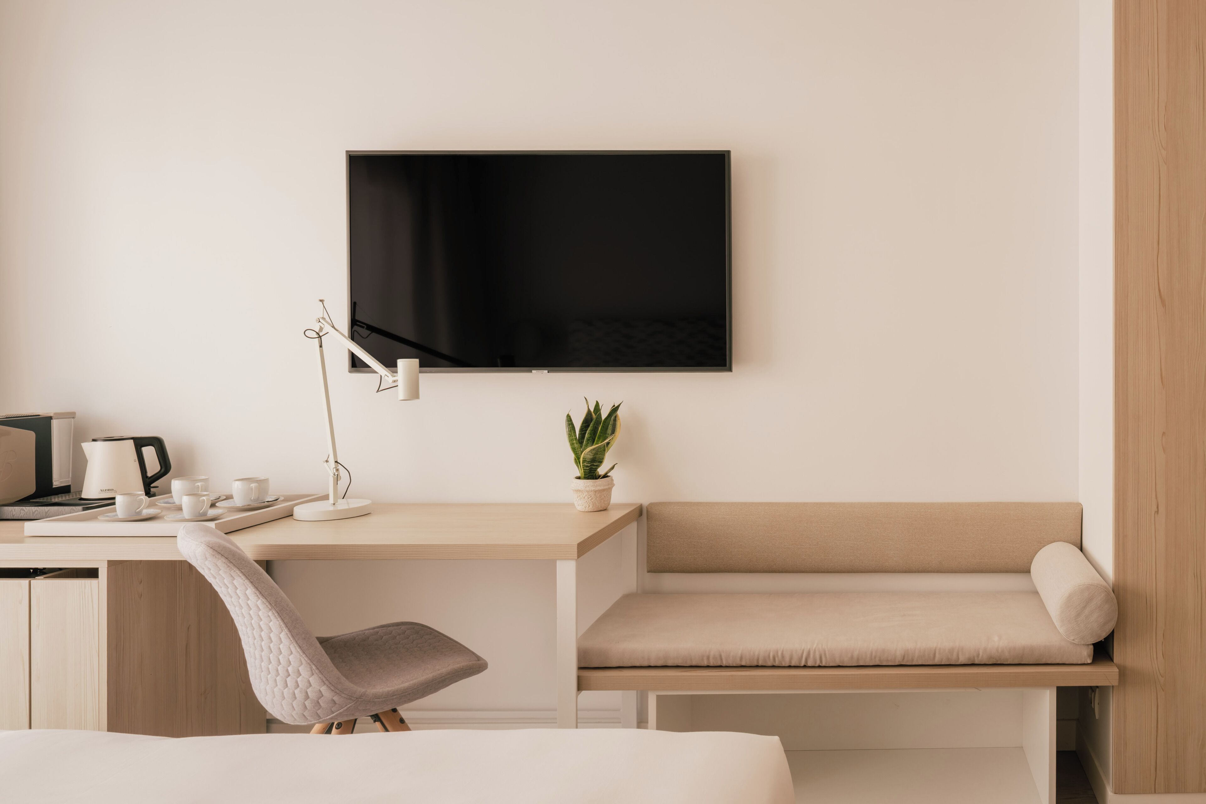 Habitación de hotel moderna con televisor de pantalla plana en la pared, un escritorio con lámpara, una silla, un sofá y una planta en maceta.