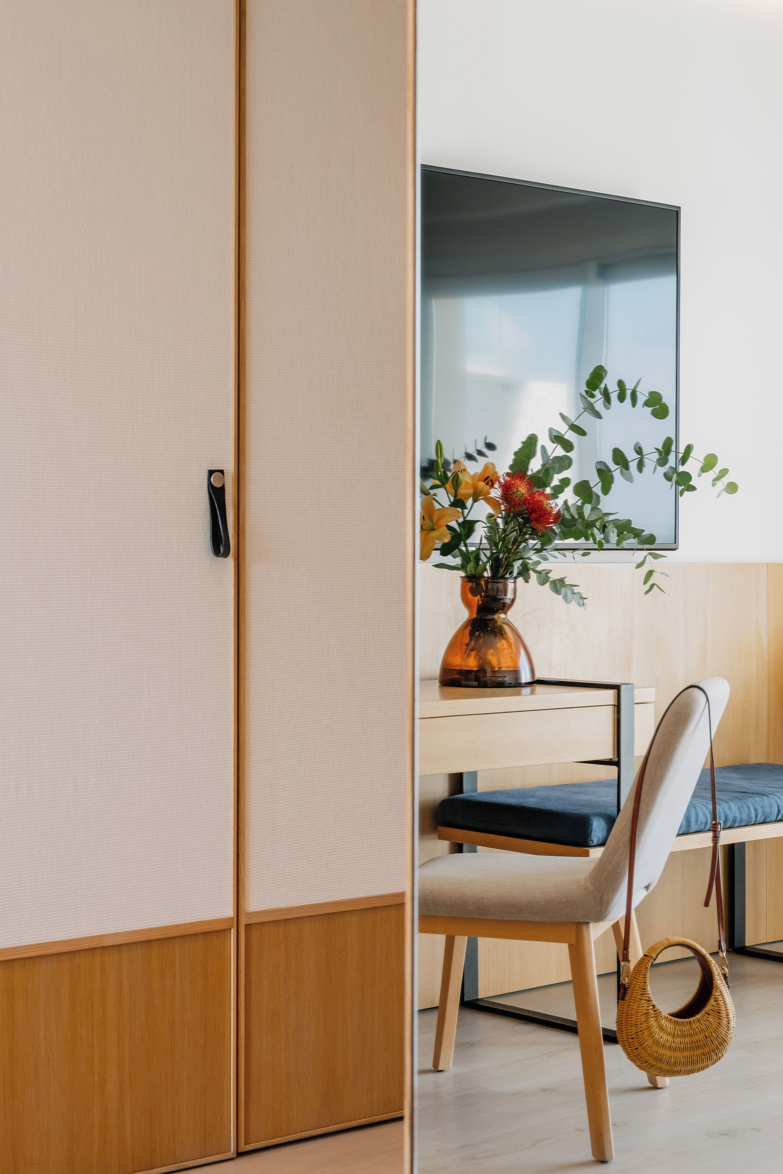 A room with a wooden wardrobe, a desk with a vase of flowers, a chair, and a television.