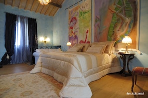 Een slaapkamer met een groot bed, een nachtkastje, een lamp, een stoel, een raam met gordijnen en een schilderij aan de muur.