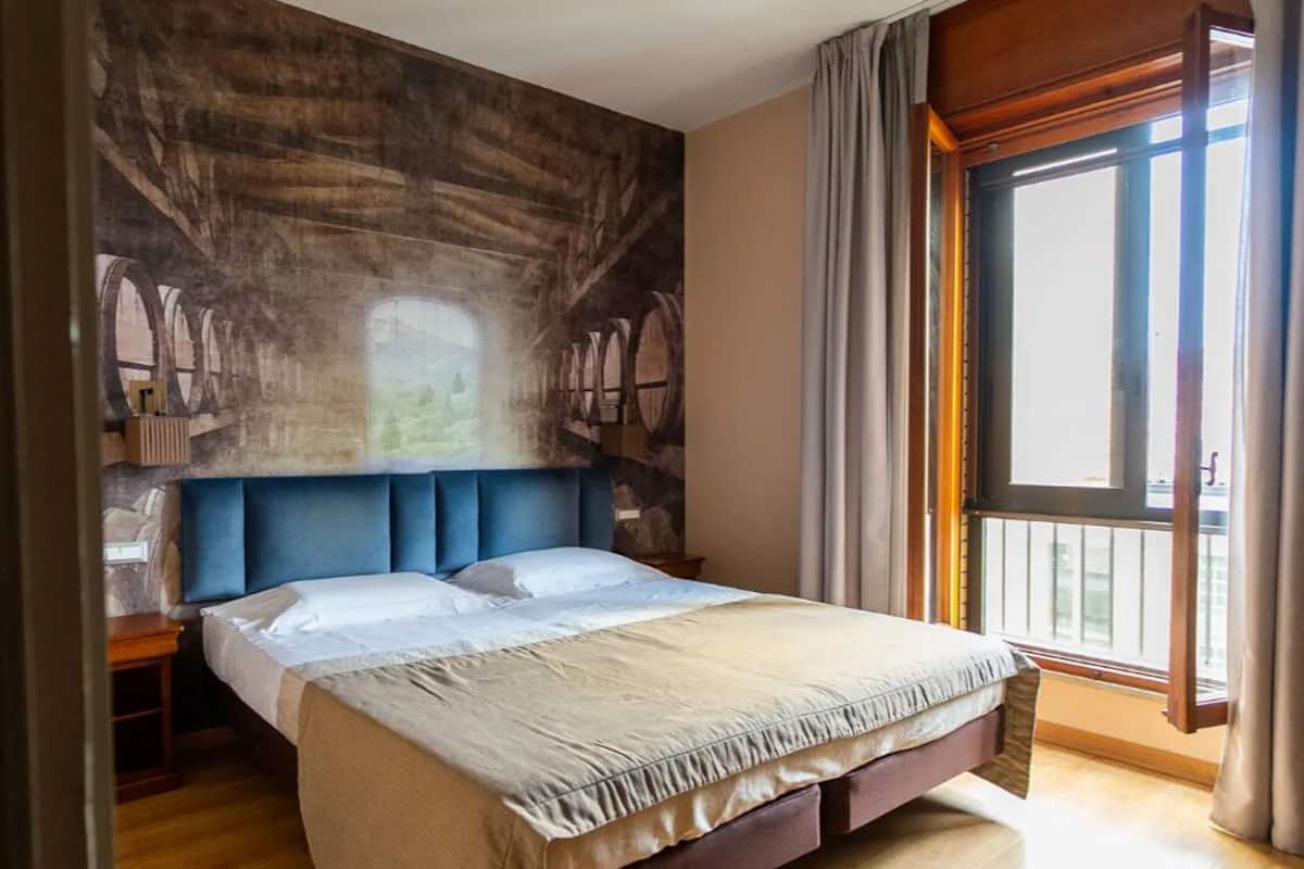 A spacious bedroom with a large bed, a wooden headboard, and a view of the outdoors through a large window.