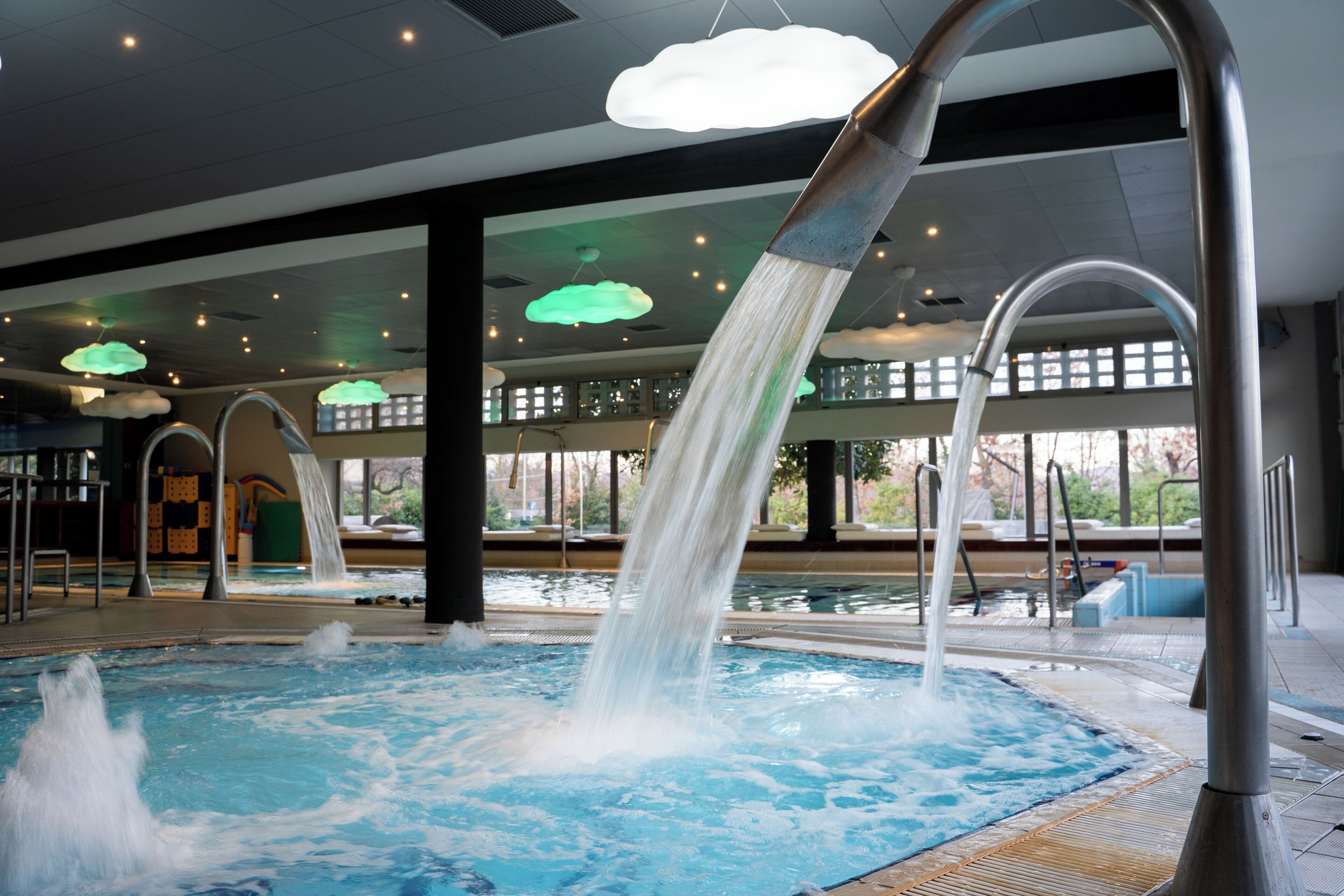 A swimming pool with multiple water jets, indoor lighting, and large windows.