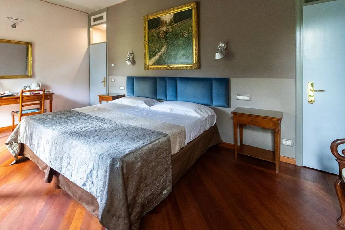 A bedroom with a large bed, a desk, and a chair. There is a mirror on the wall and a door to the right.