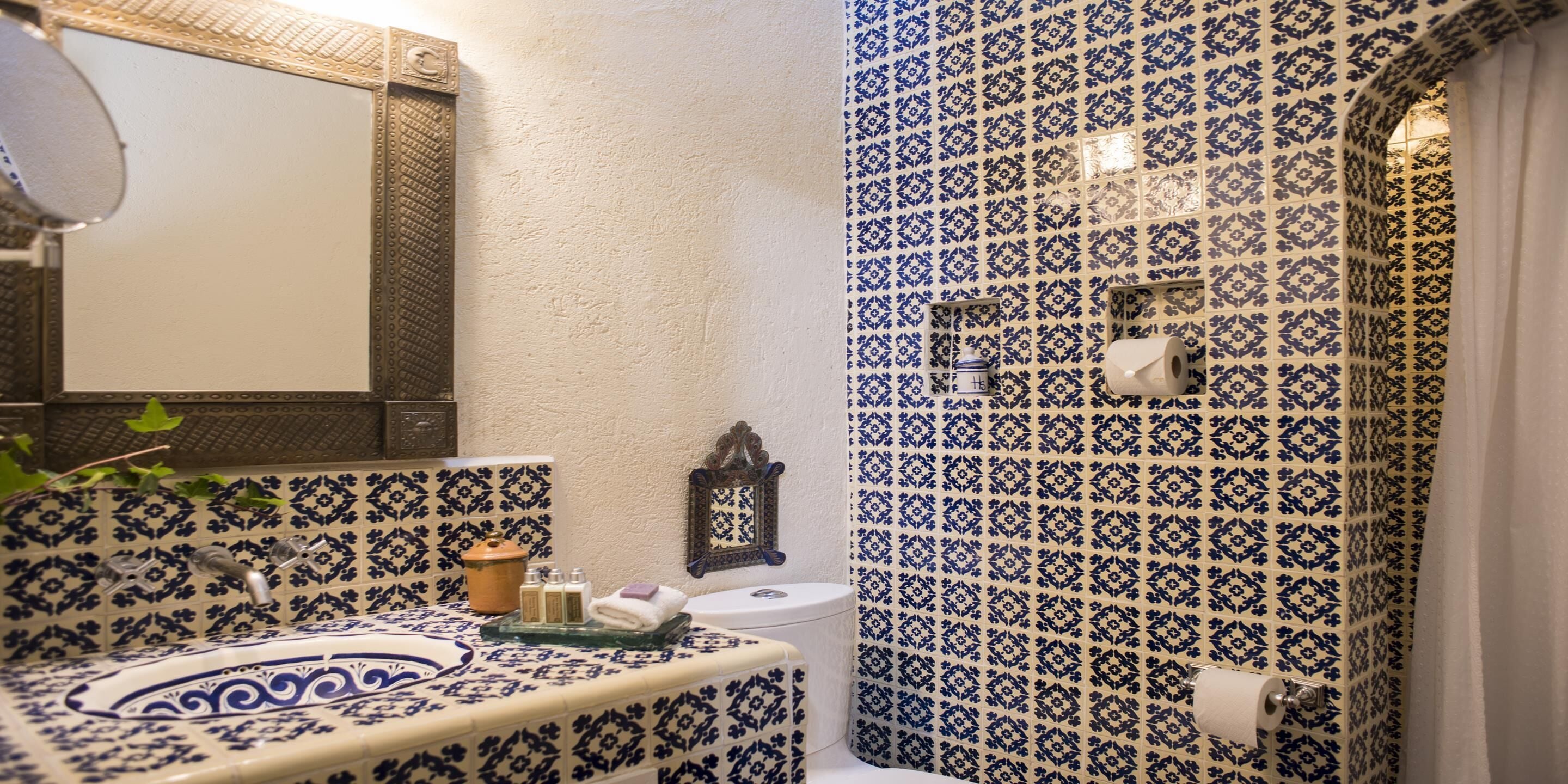Une salle de bain avec des carreaux bleus et blancs formant un motif, un lavabo avec un récipient décoratif, un miroir et des toilettes avec un rouleau de papier toilette.