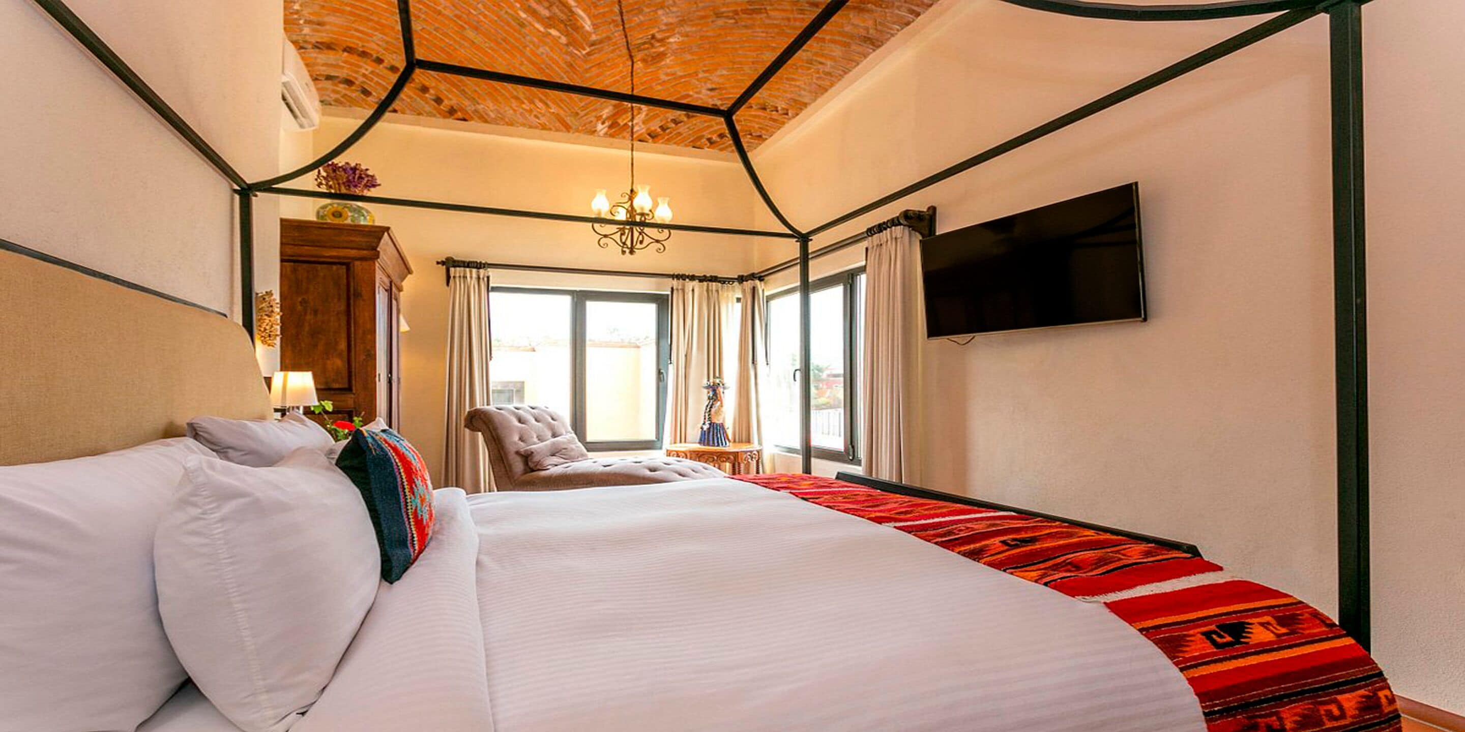 A hotel room with a four-poster bed, a flat-screen TV mounted on the wall, and a large window with curtains.