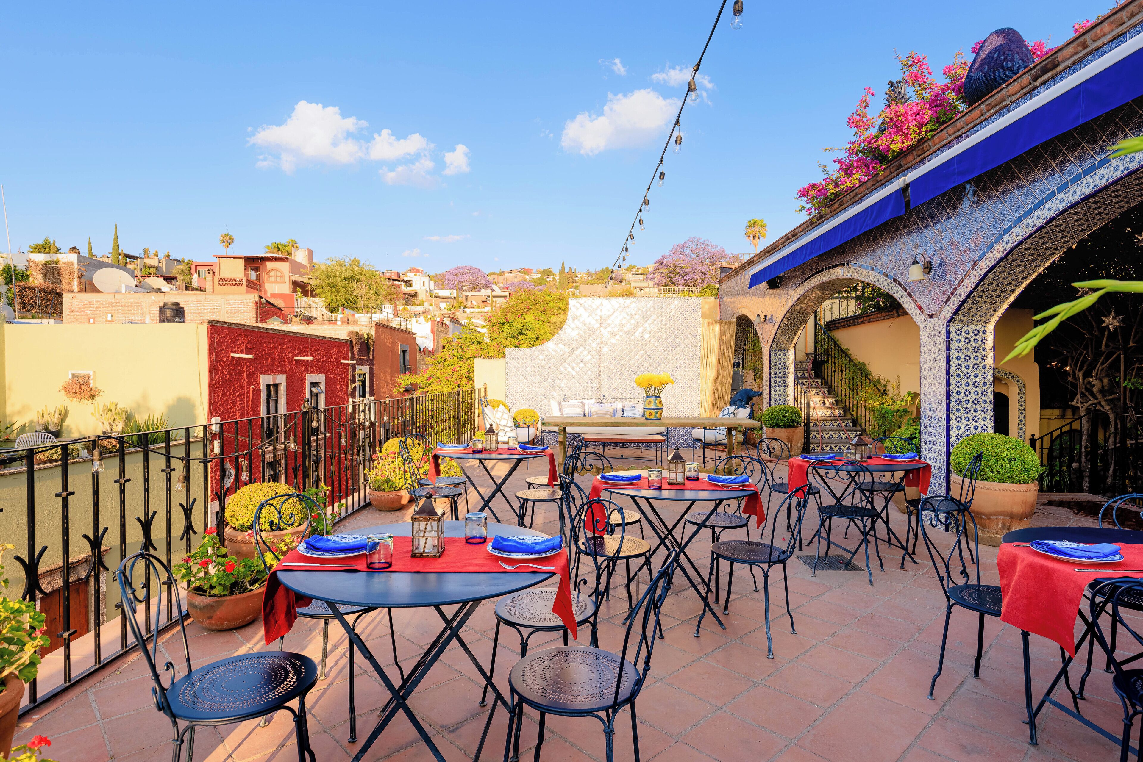 Un patio en la azotea con mesas y sillas, rodeado de edificios y un arco decorativo.