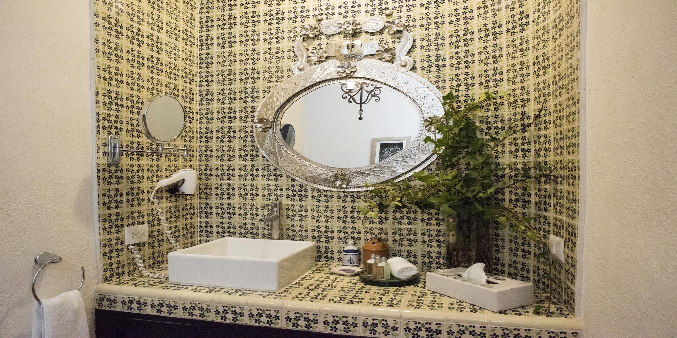 A bathroom with a floral tile backsplash, a round mirror, a rectangular sink, and a small plant.