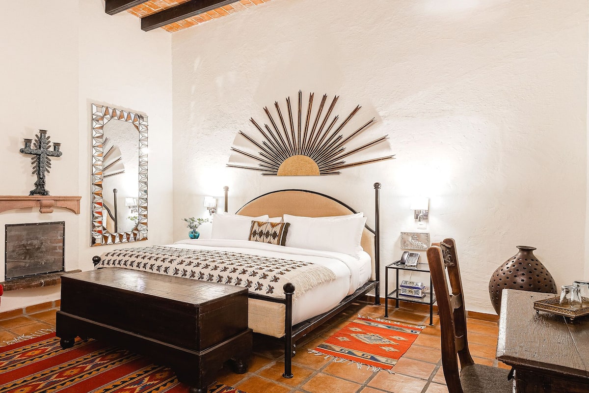 A bedroom with a bed, a red armchair, a wooden coffee table, a patterned rug, and a decorative mirror.