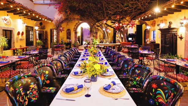Una larga mesa de comedor con sillas, un arreglo floral central y un balcón con plantas e iluminación.