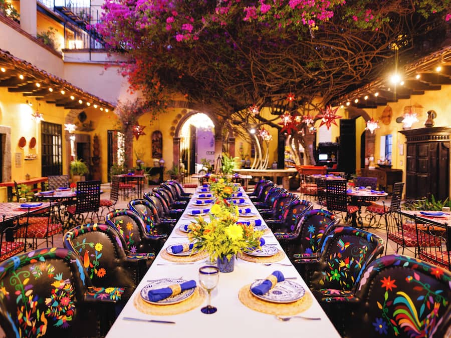 A long dining table with chairs, a central floral arrangement, and a balcony with plants and lights.