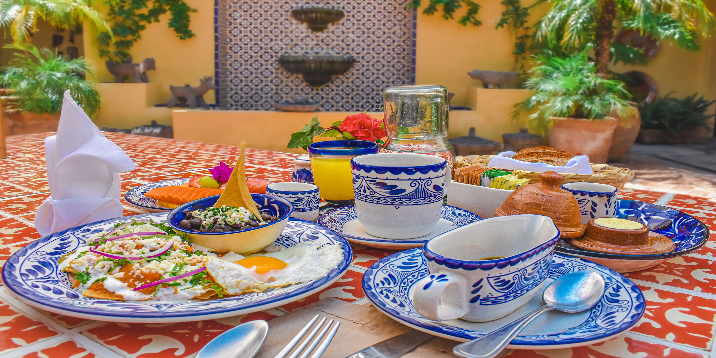 Una mesa preparada para el desayuno, con huevos, tostadas y un tazón de frijoles, platos y tazas con diseño azul y blanco.