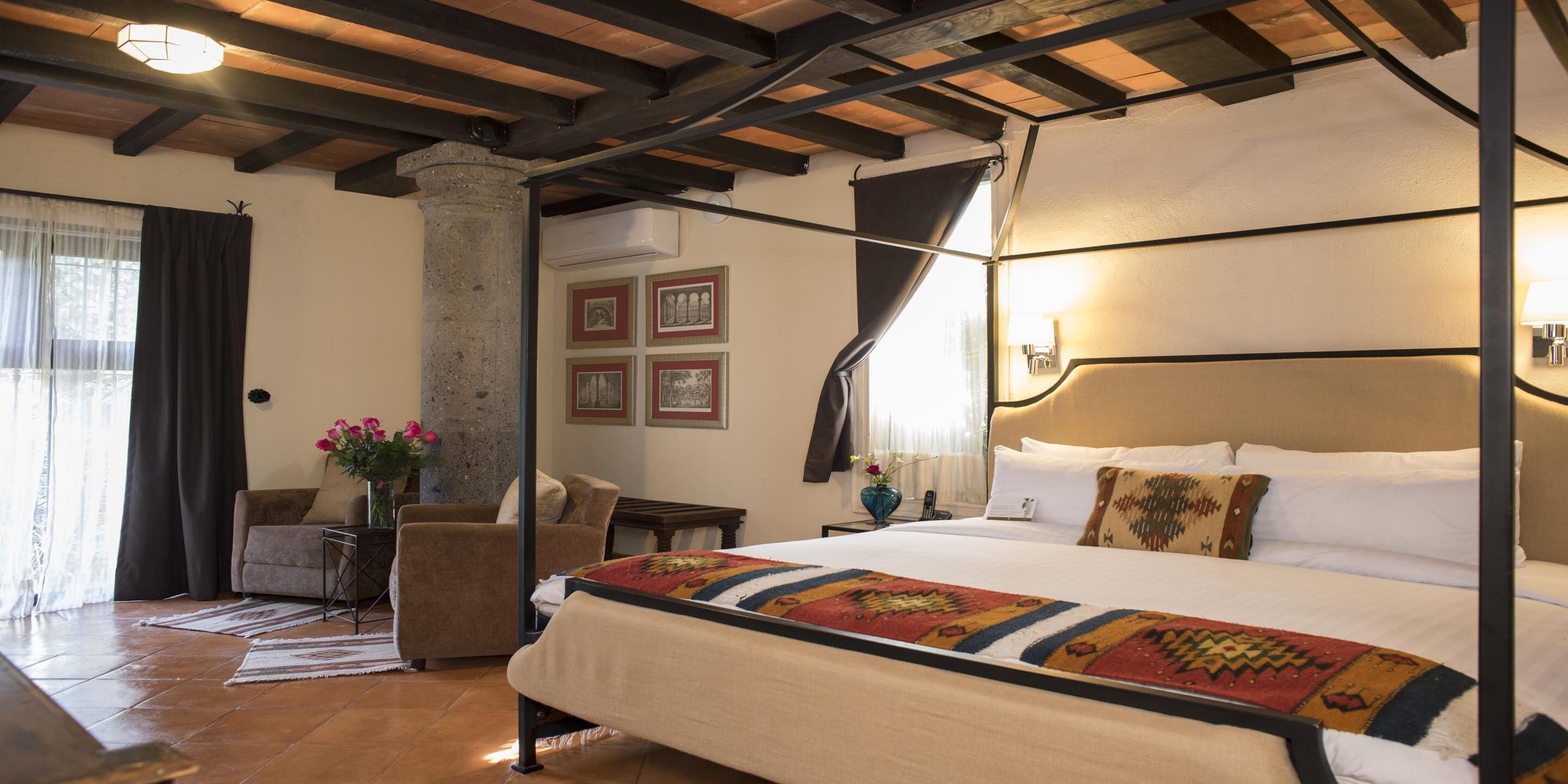A four-poster bed with a colorful blanket, a stone pillar, a sofa, and a small table with a vase of flowers.