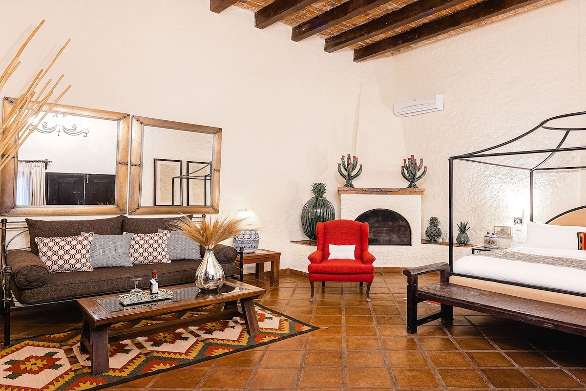 A room with a bed, a sofa, a red armchair, a coffee table, and a fireplace.