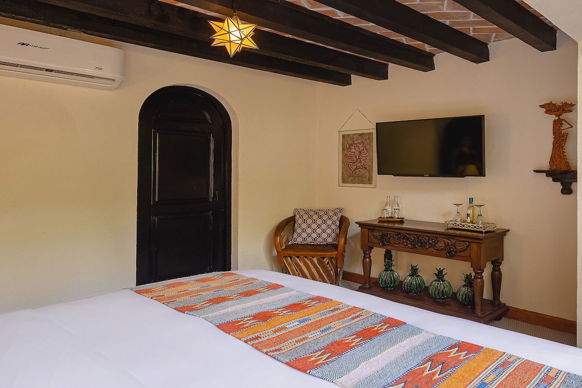 A bedroom with a bed, a TV mounted on the wall, a wooden chair, a small table with decorative items, and a door.