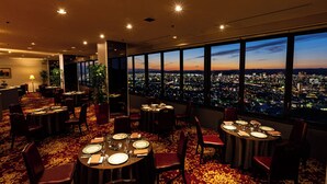 A hotel dining area with tables set for dinner, large windows offering a city view, and a carpeted floor. - Okayama International Hotel (Okayama)