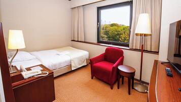 A hotel room with a bed, a red armchair, a small table, a desk with a TV, and a window with curtains.