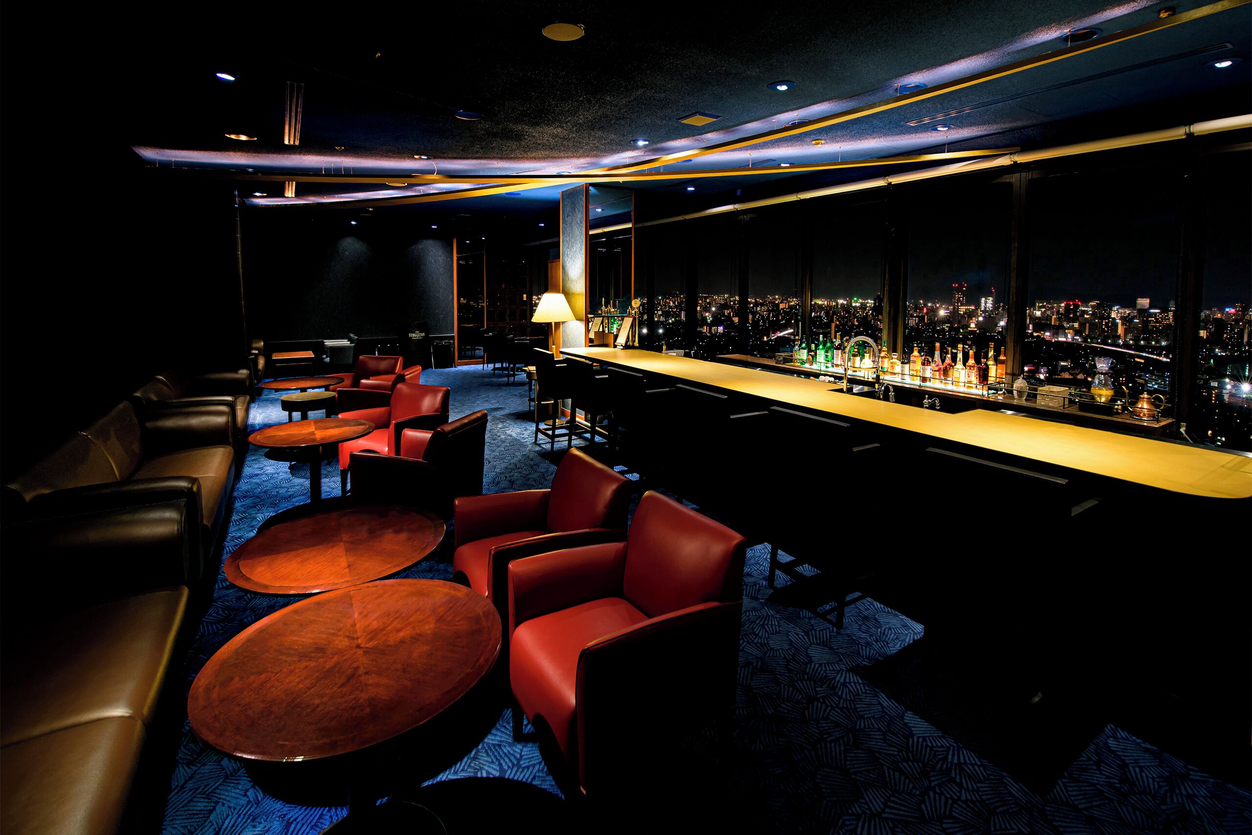 A bar area with leather seating, a long counter with bottles, and a city view through large windows.