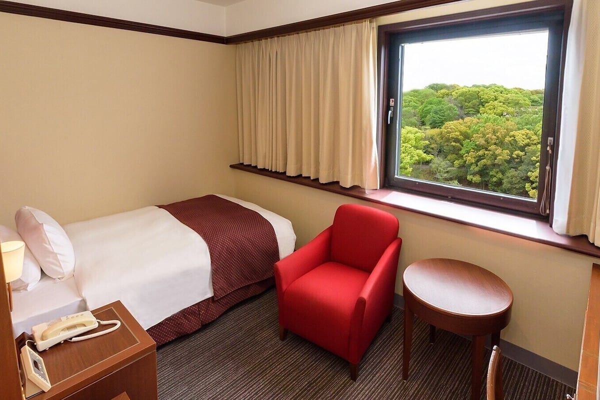 Een hotelkamer met een bed, een rode stoel, een tafeltje, een telefoon en een raam met gordijnen.