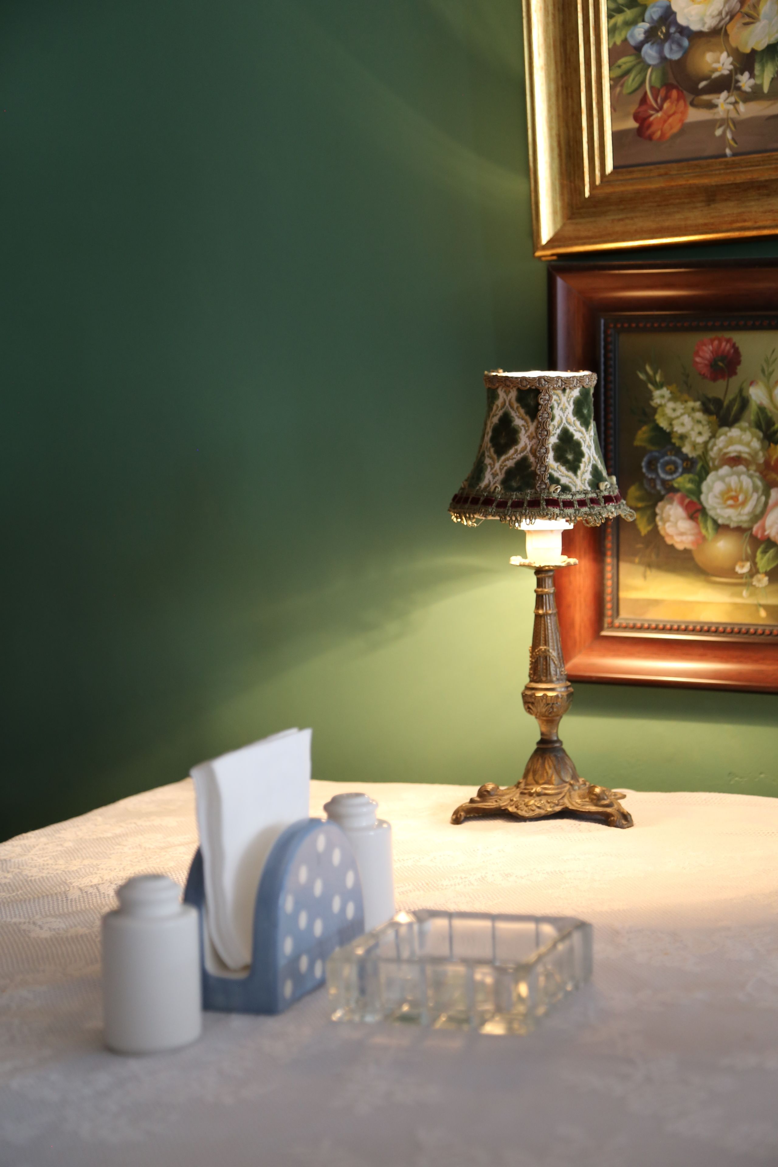 A table with a decorative lamp, a tissue box, and a small container, against a green wall with a framed floral painting.