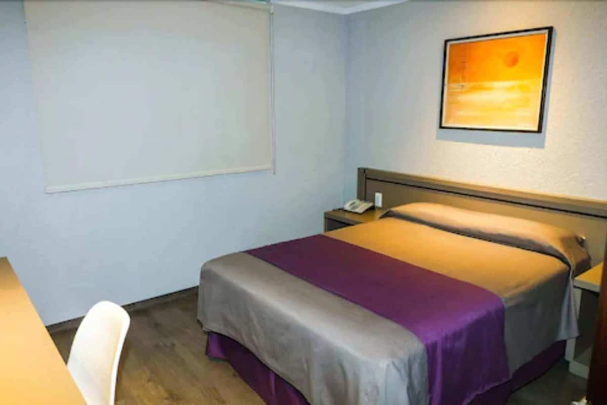 A single bed with a headboard, a bedside table with a phone, a chair, and a window with blinds.