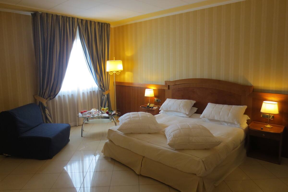 A hotel room with a large bed, two bedside lamps, a small table with a tray of breakfast items, a blue armchair, and a window with curtains.