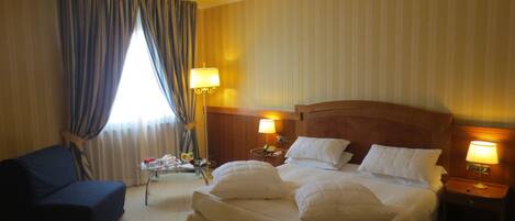 Une chambre d’hôtel avec un grand lit, deux lampes de chevet, une petite table sur laquelle se trouve un plateau avec des articles pour le petit-déjeuner, un fauteuil bleu et une fenêtre avec des rideaux.