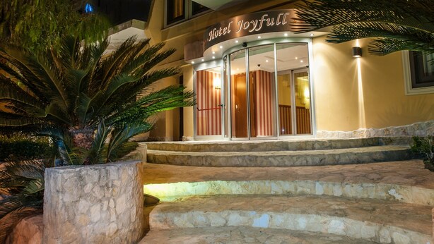 Entrada de hotel con un letrero que dice "Hotel Joyfull", que presenta una puerta de cristal, escalones de piedra y una palmera en primer plano.