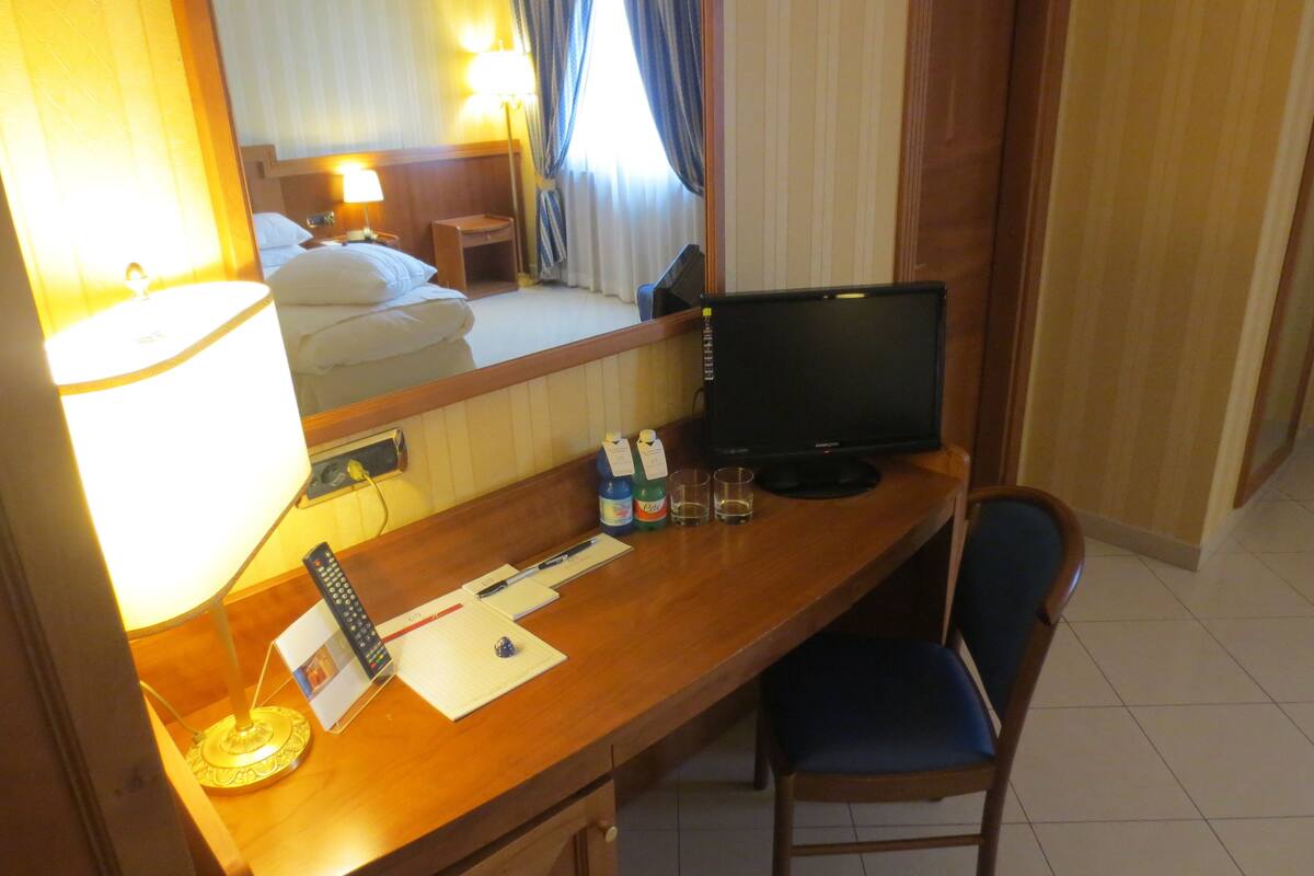 A hotel room with a wooden desk, a computer monitor, a lamp, a chair, and a bed with white linens.