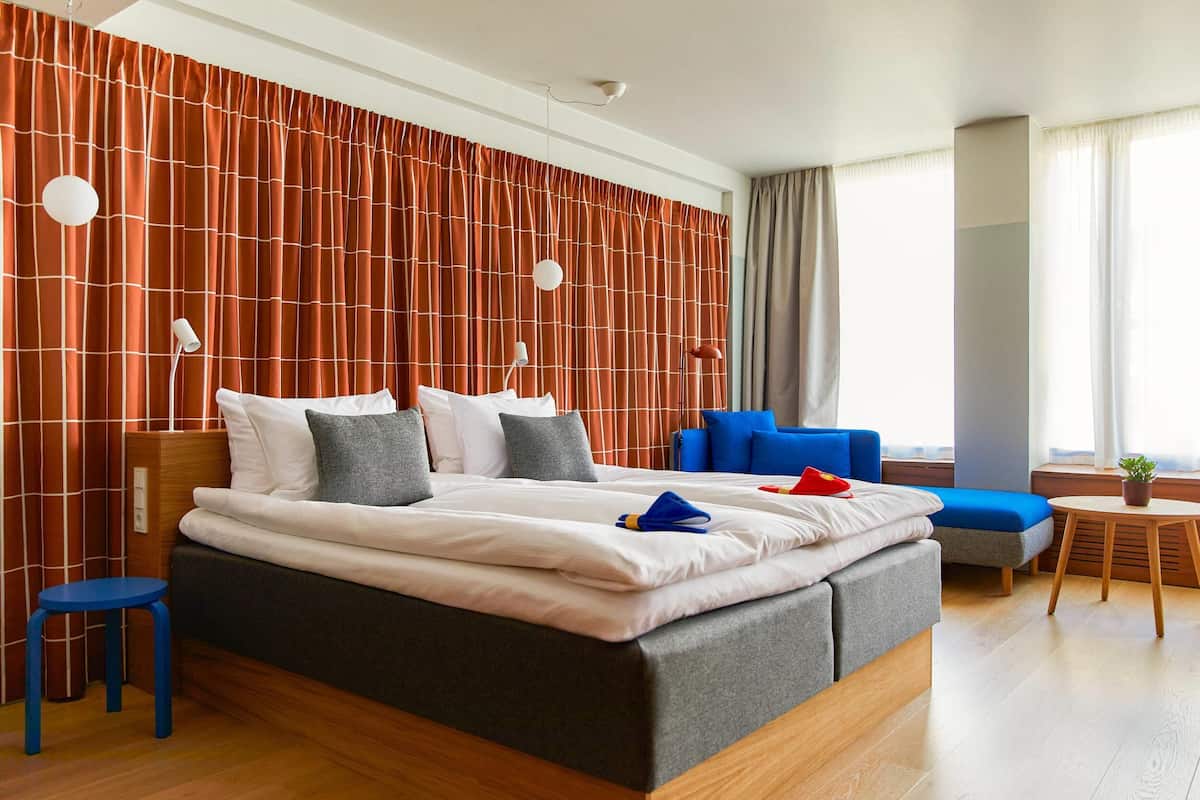 A modern hotel room with a large bed, a blue sofa, a small round table, and a view of the city.