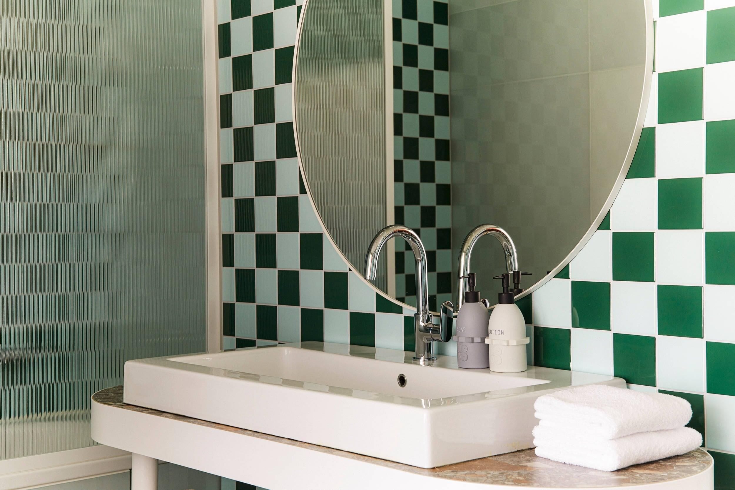 A bathroom with a white sink, two faucets, a round mirror, and a green and white checkered tile wall.
