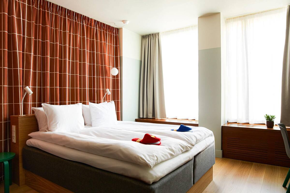 A hotel room with a large bed, a desk, a chair, and a window with curtains.