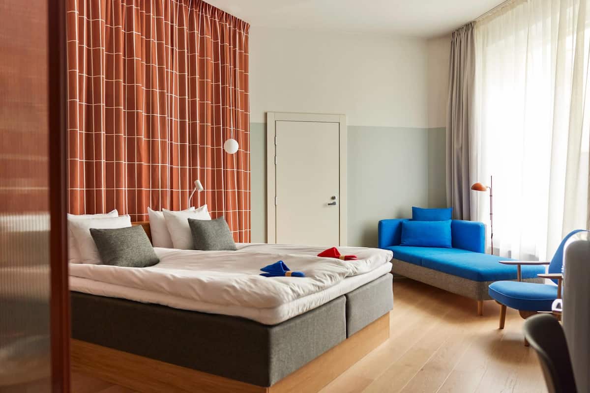 A modern hotel room with a large bed, a sofa, and a chair. There is a door and a window with curtains.