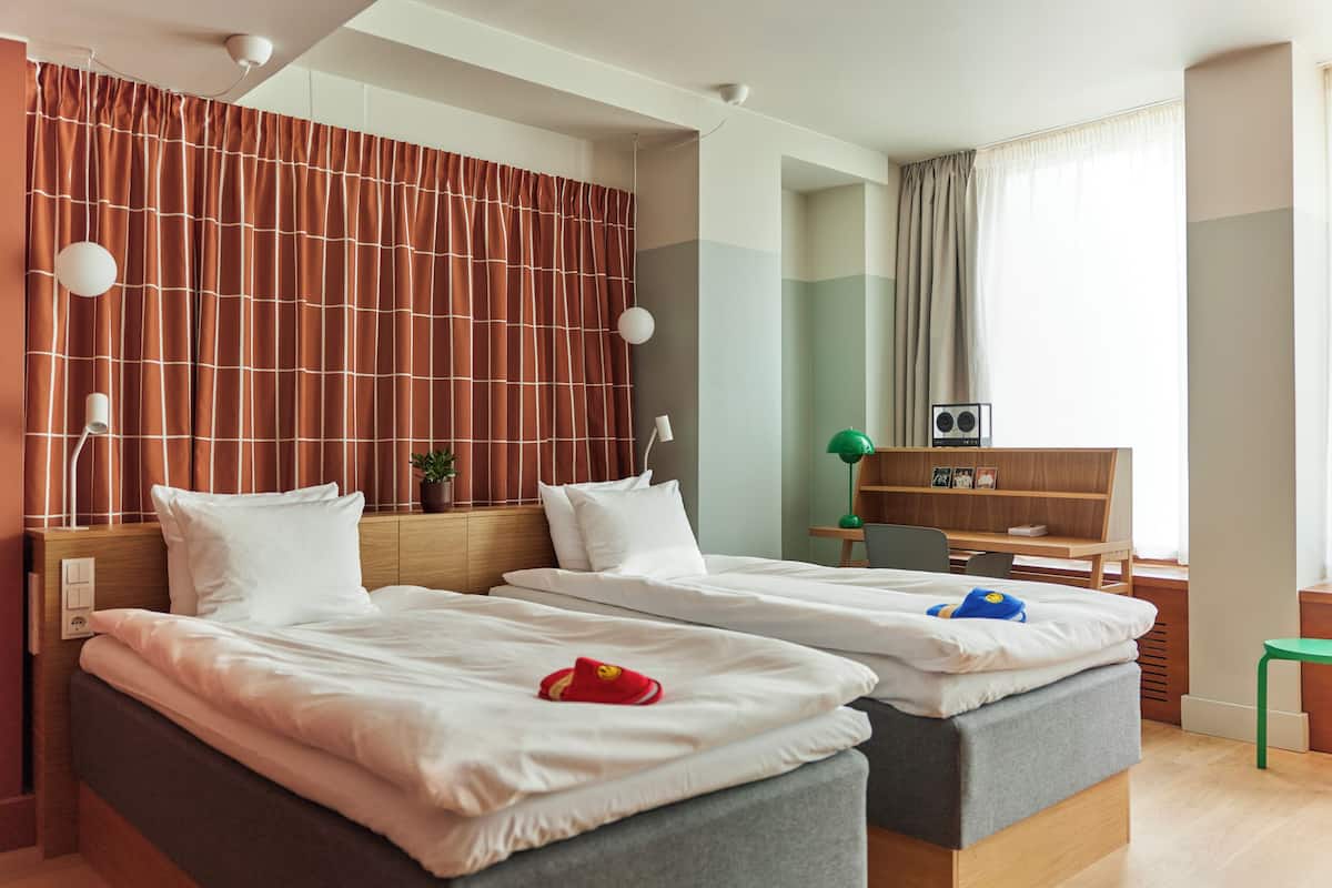 A hotel room with two beds, a desk, a chair, a TV, and a window with curtains.