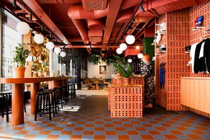 A modern cafe with a checkered floor, wooden tables, black stools, and a brick-patterned wall with hanging plants and a mannequin. - Hobo Helsinki (Helsinki)