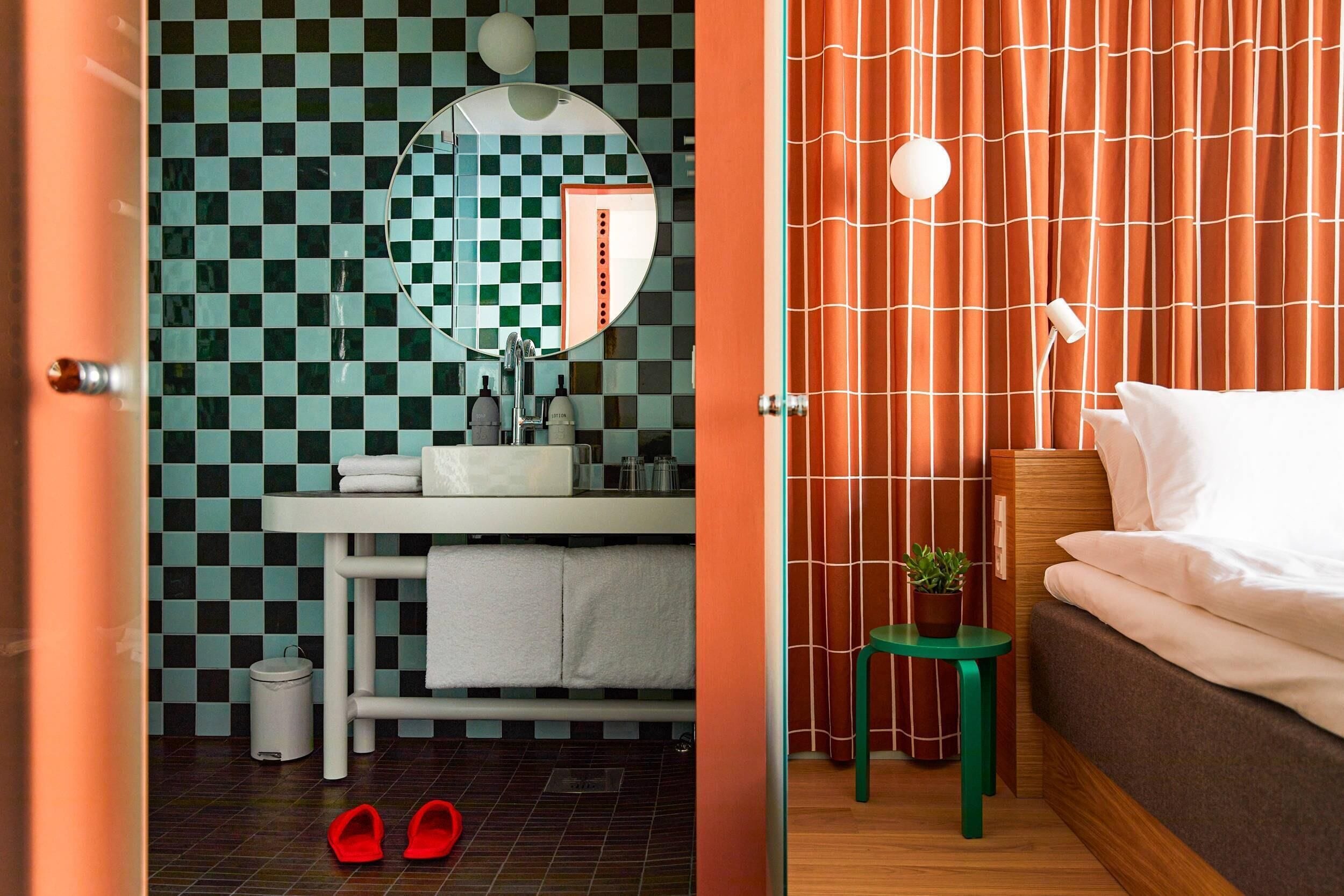 Un baño con pared de baldosas a cuadros, un lavamanos y un espejo. Un par de pantuflas rojas están en el suelo.