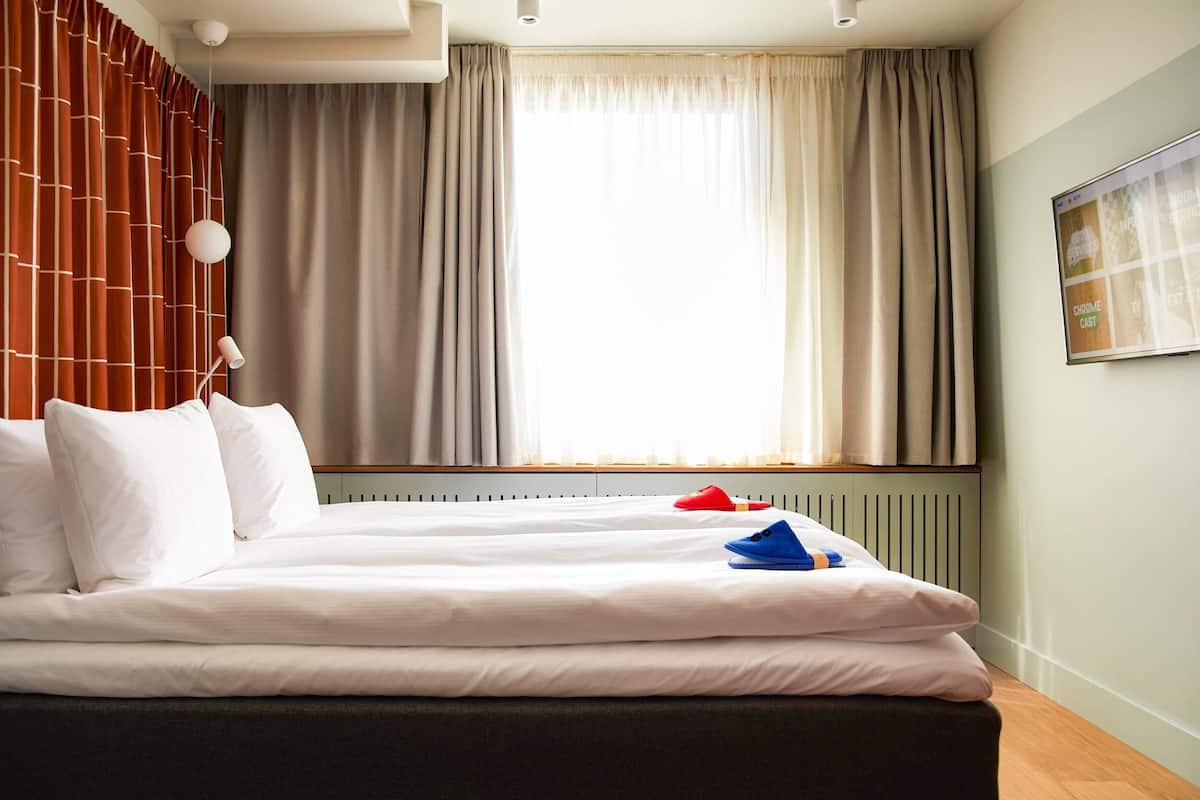 A hotel room with a large bed, a window with curtains, a wall-mounted TV, and a radiator.