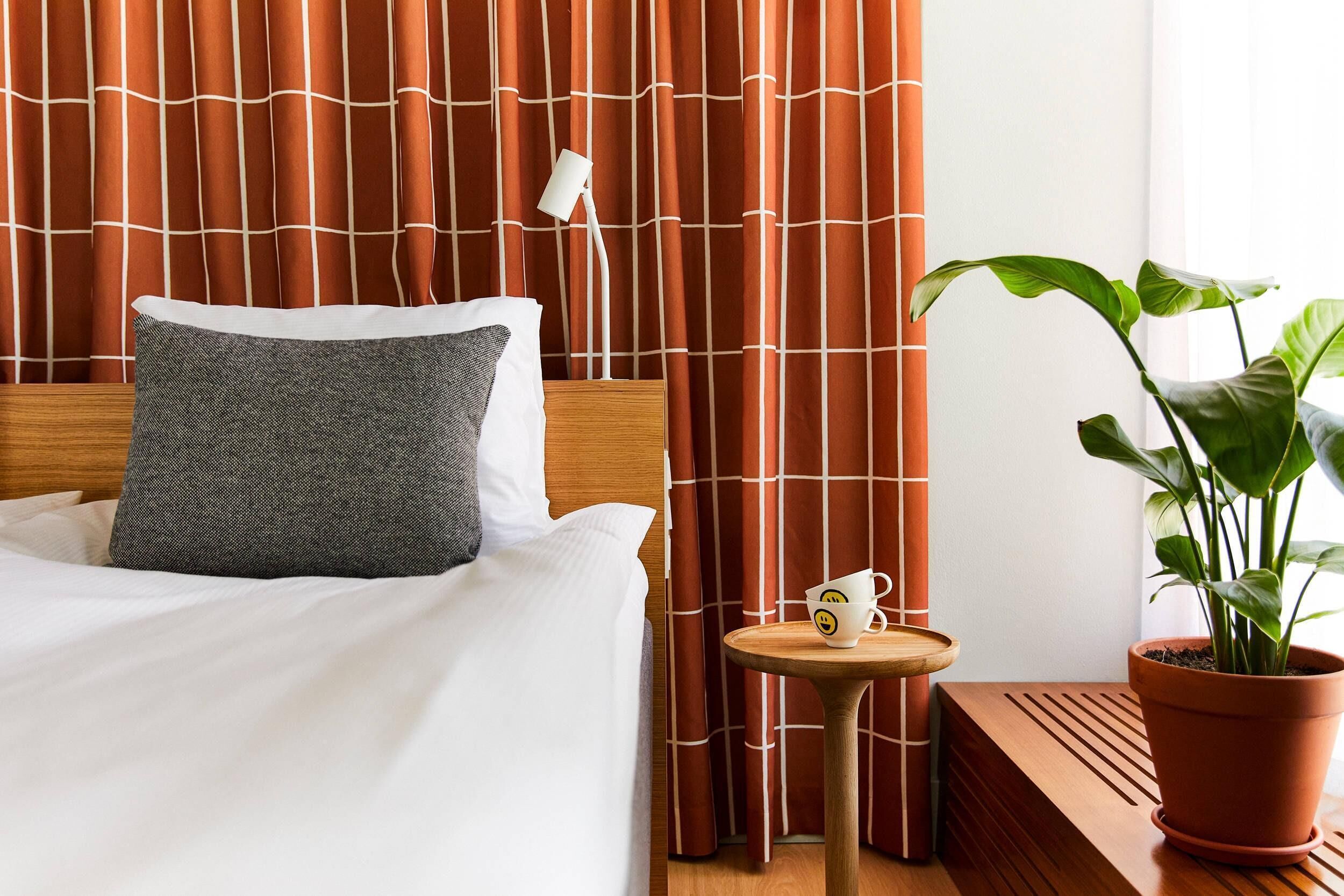 A neatly made bed with a grey pillow, a small table with a mug, and a potted plant.