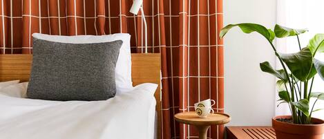 A neatly made bed with a grey pillow, a small table with a mug, and a potted plant.