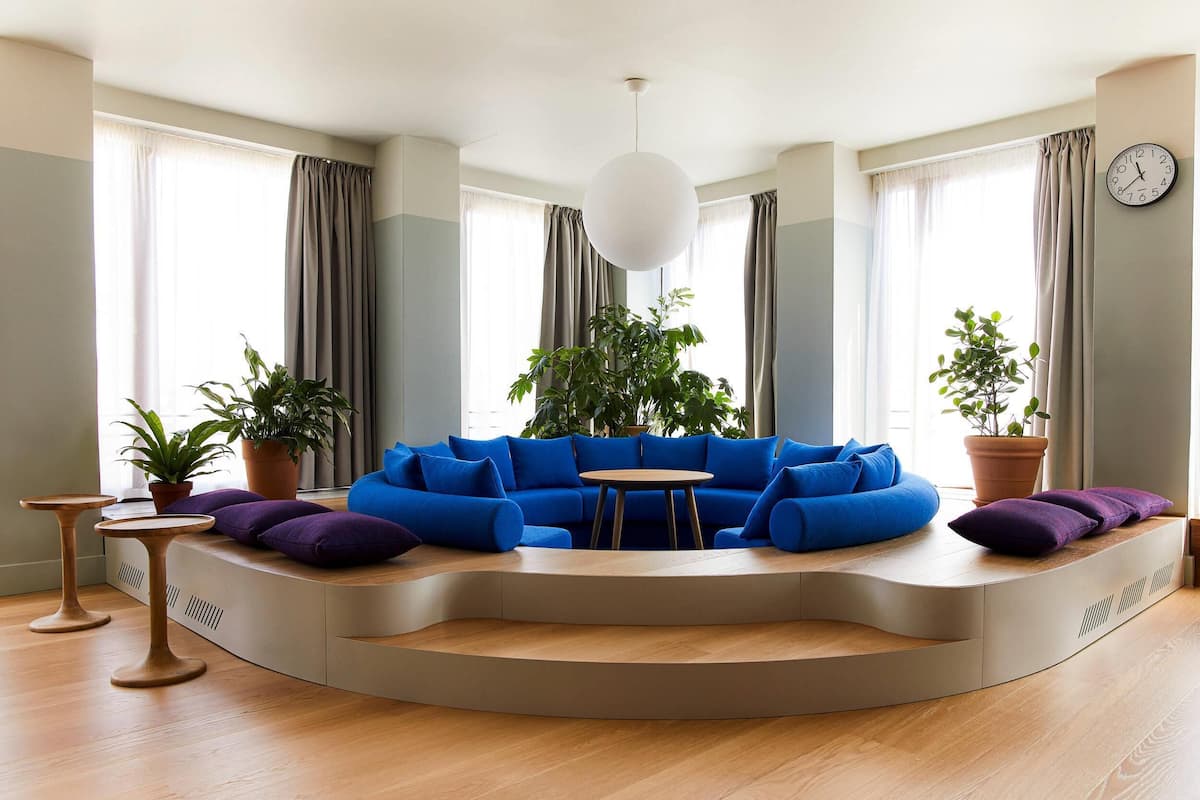 A modern living room with a blue sofa, a round coffee table, and two side tables. There are potted plants and a large window with curtains.