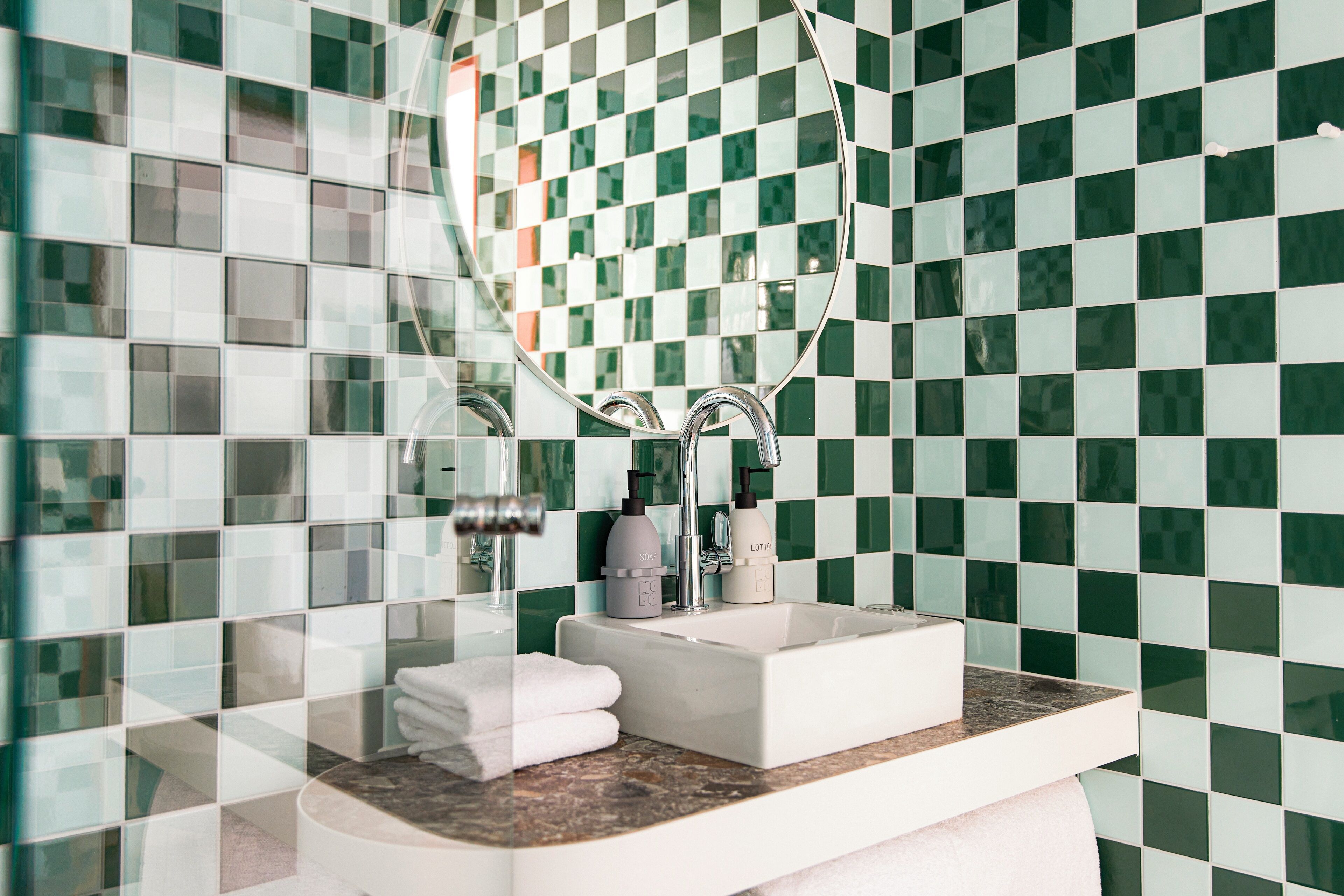 A bathroom with a checkered green and white tile backsplash, a white sink with a marble countertop, and a round mirror.