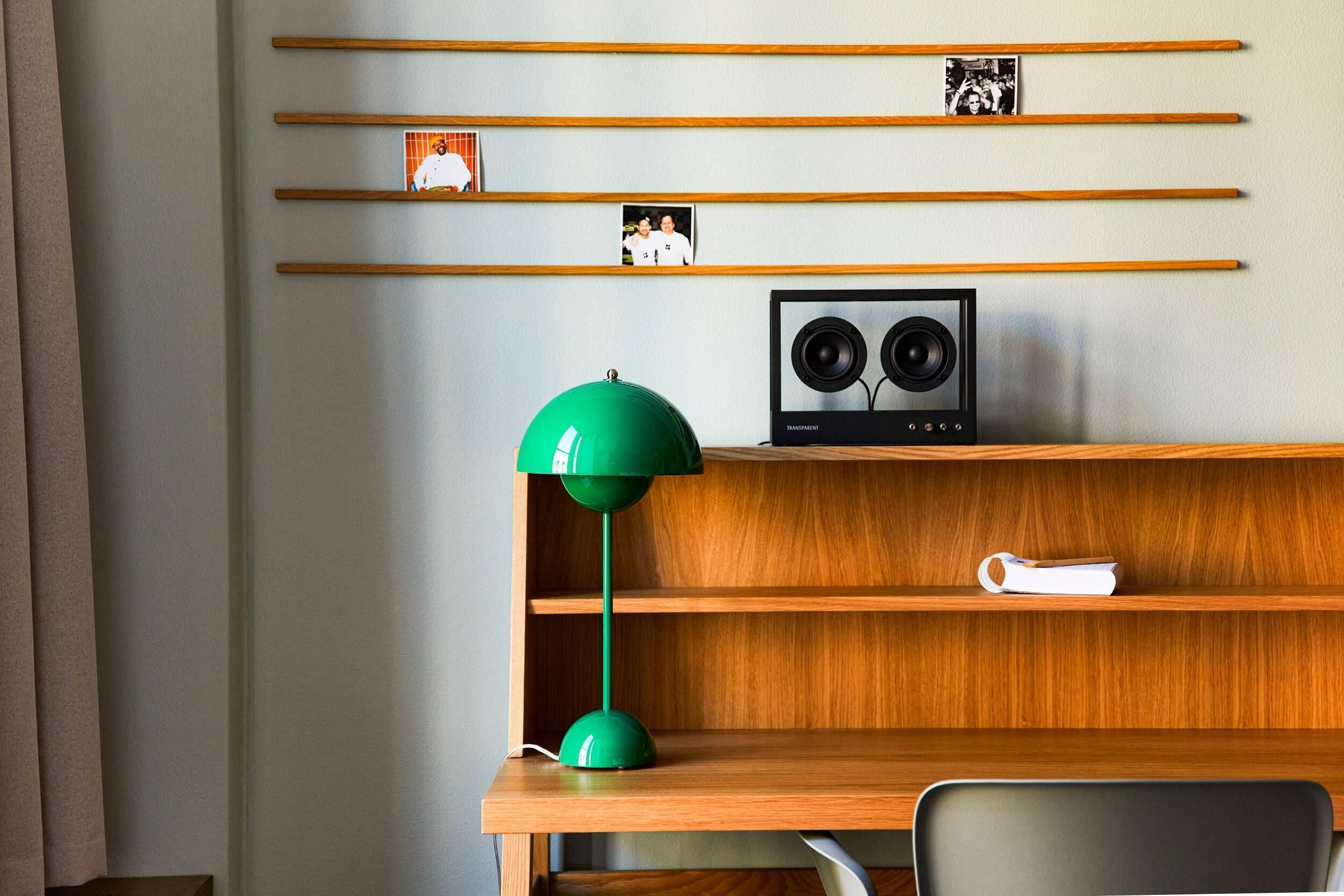Una stanza con una scrivania in legno, una lampada da tavolo verde, una sedia bianca, uno scaffale con fotografie e un altoparlante.