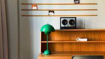A room with a wooden desk, a green desk lamp, a white chair, a shelf with photos, and a speaker.
