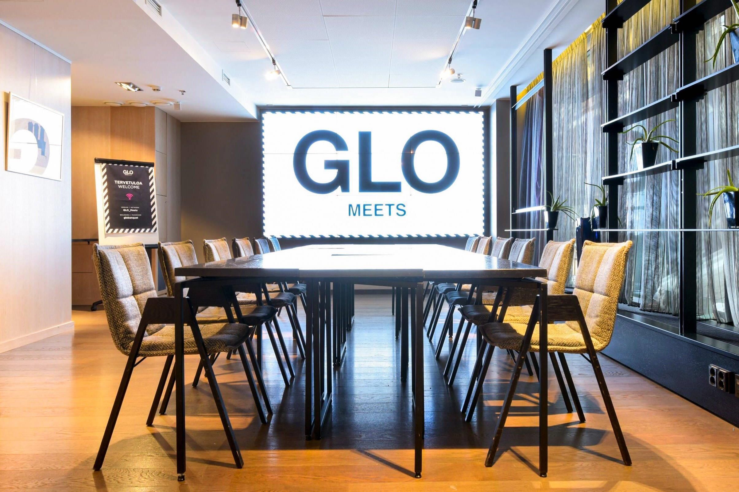 Un salón de conferencias con una gran mesa, sillas, una pantalla mostrando 'GLO MEETS' y una estantería con plantas.