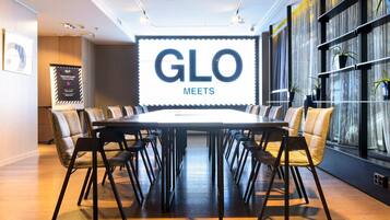 Un salón de conferencias con una gran mesa, sillas, una pantalla mostrando 'GLO MEETS' y una estantería con plantas.