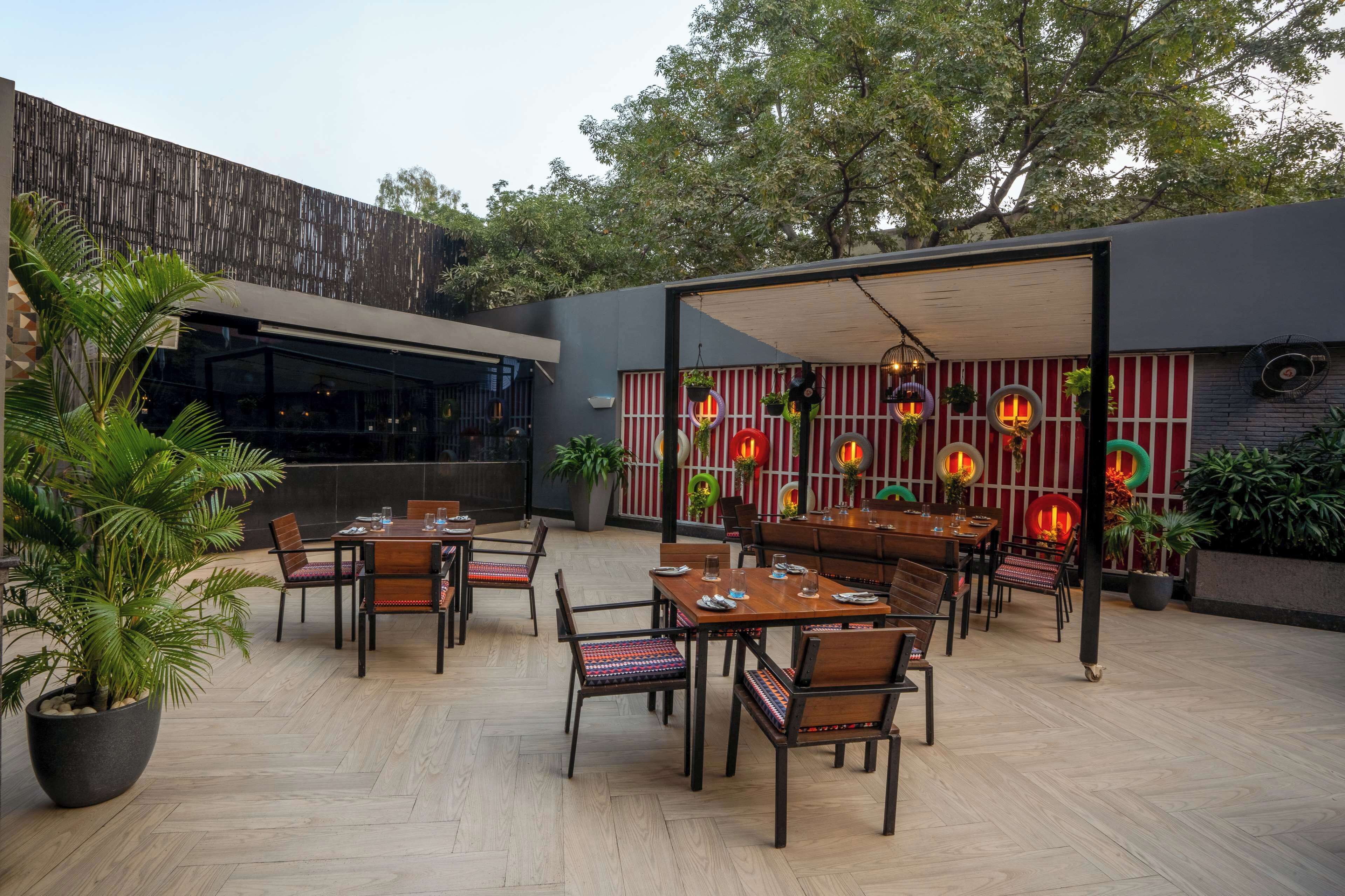 Un área de comedor al aire libre con varias mesas y sillas, rodeada de plantas en macetas y una fachada de edificio moderna.