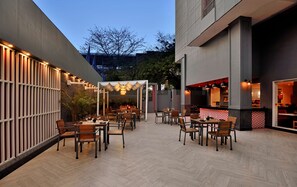 An outdoor dining area with multiple tables and chairs, a pergola with string lights, and a restaurant visible through an open doorway.