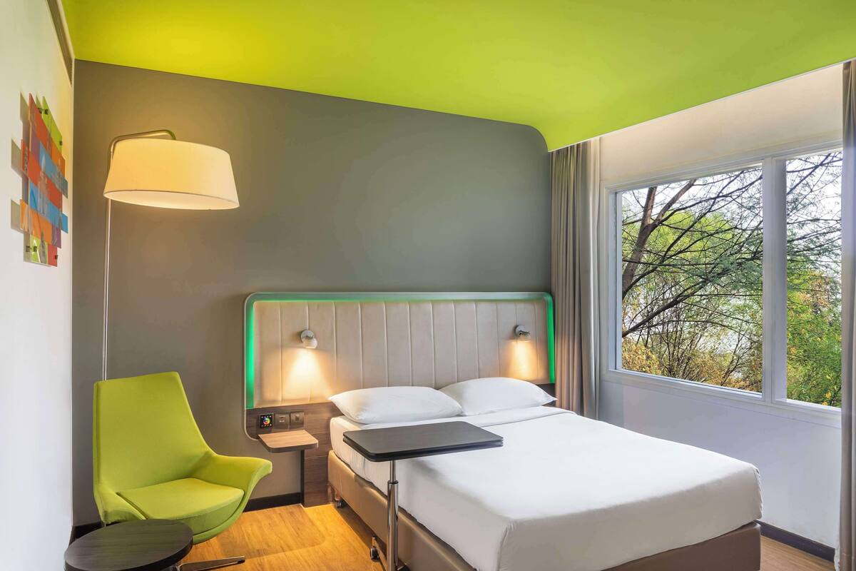 A hotel room with a bed, a green armchair, a bedside table, a lamp, and a window with a view of trees.