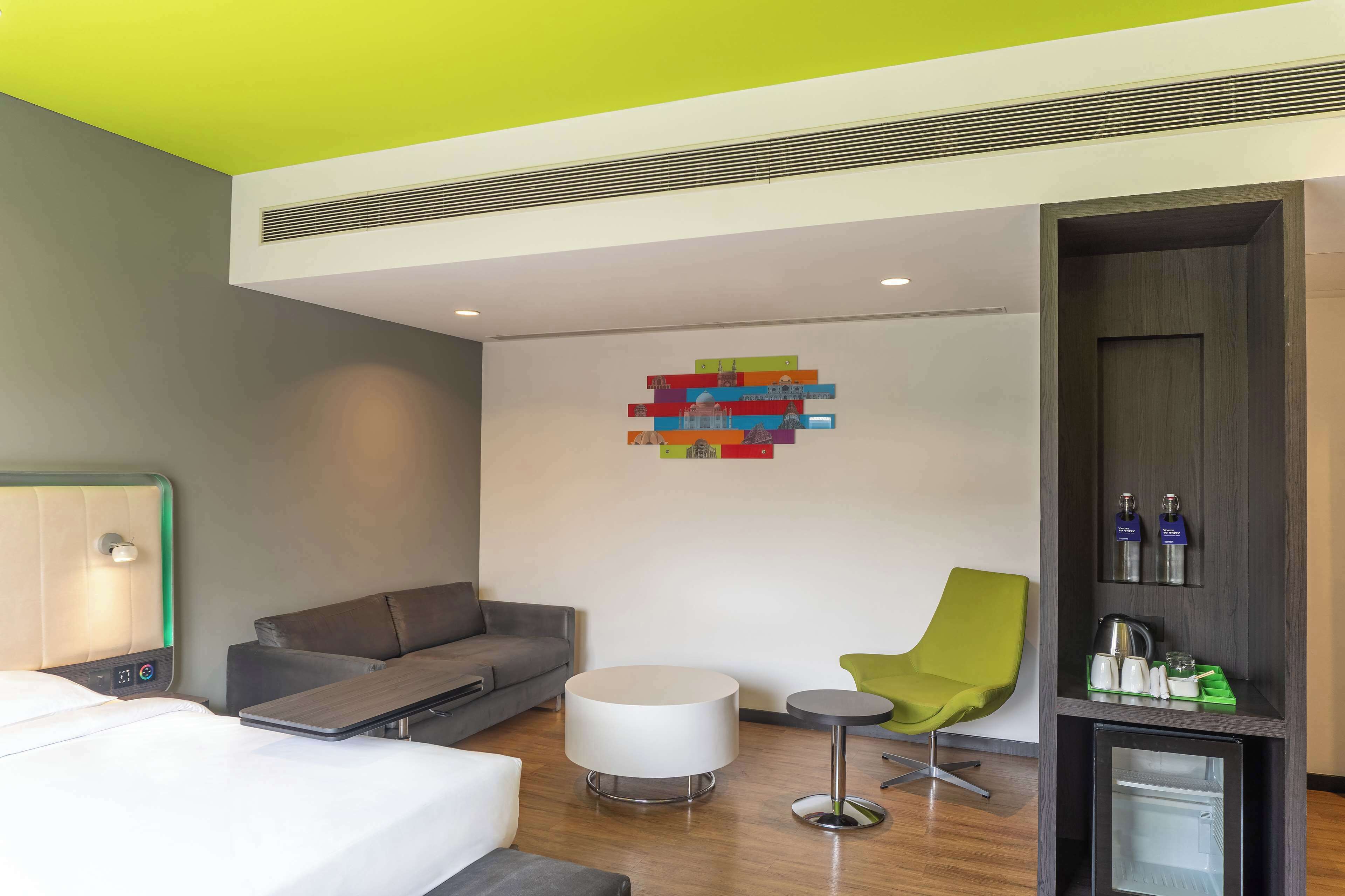 A modern hotel room with a bed, a grey sofa, a white round table, a green armchair, and a small fridge.