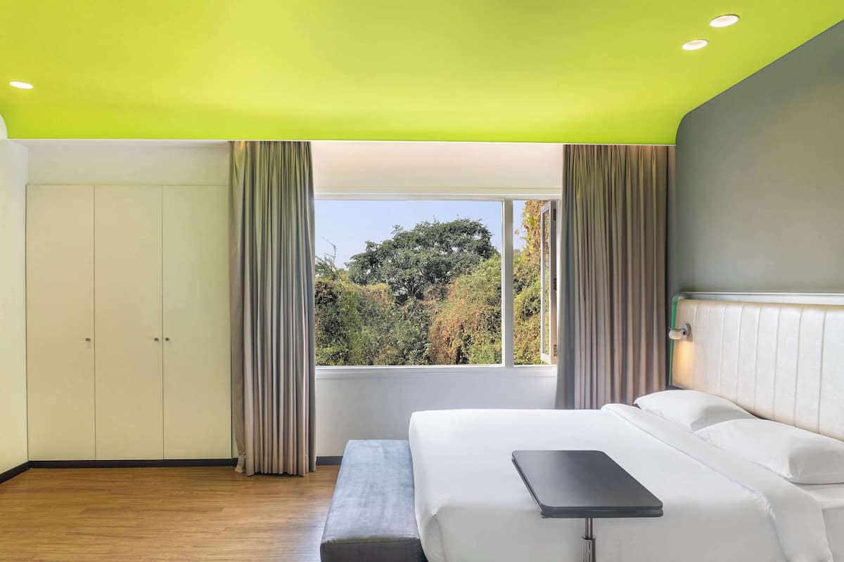 A modern hotel room with a bed, a bench, a small table, and a view of trees through a large window.