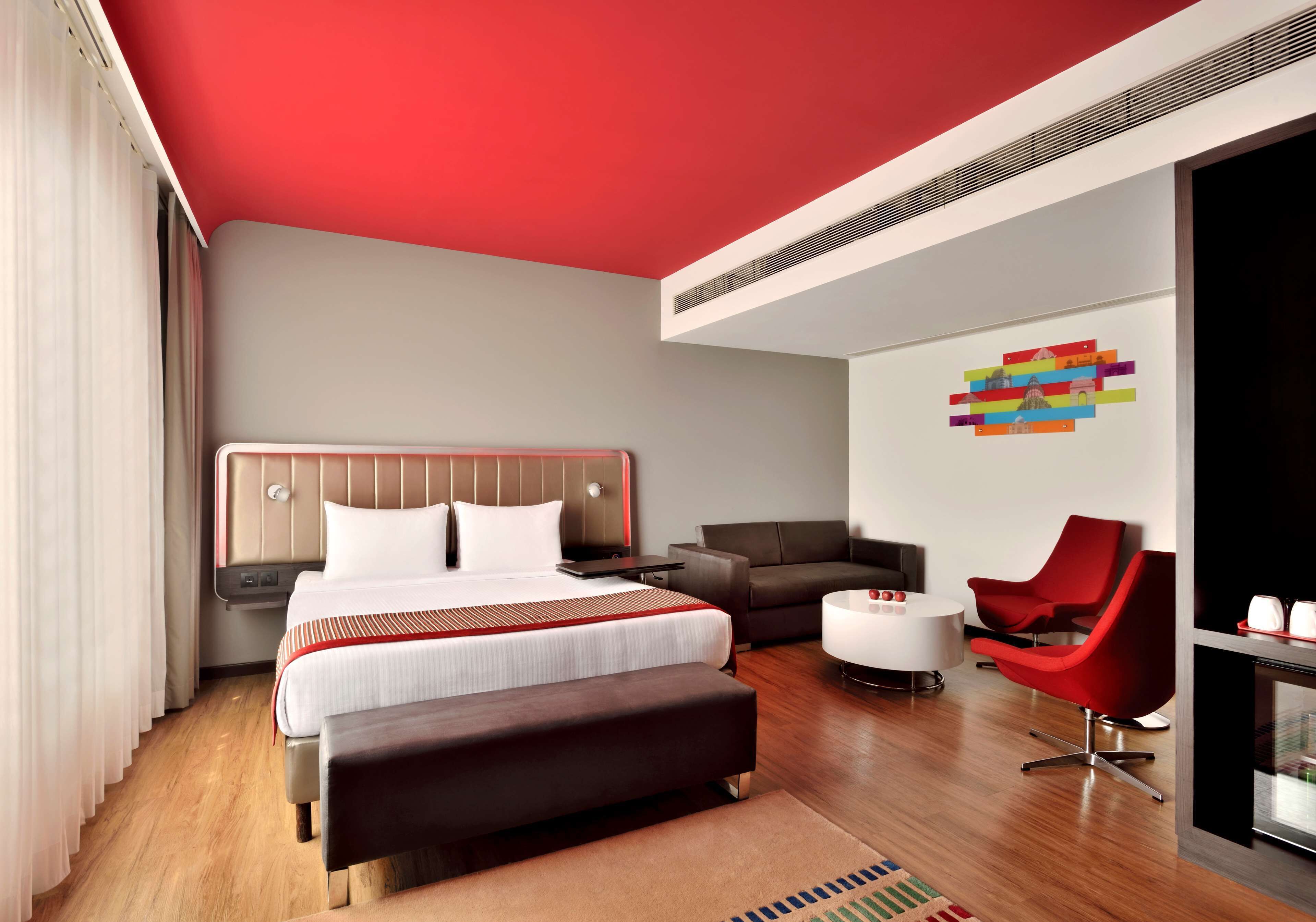 A modern hotel room with a large bed, a sofa, a red chair, and a small table.